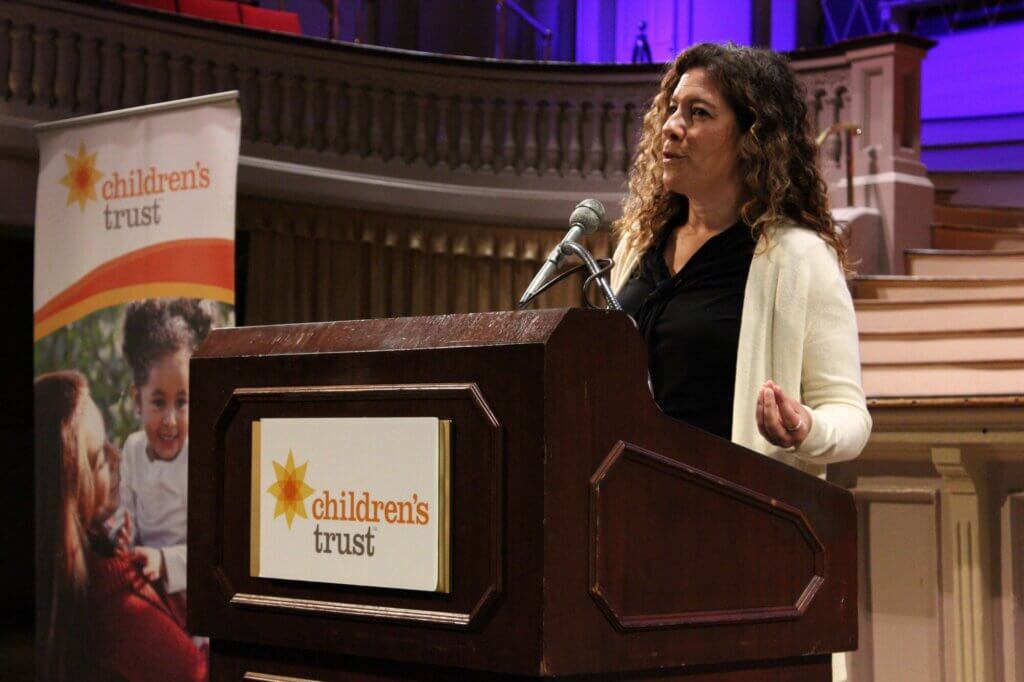 A woman with curly hair speaks at a podium with a Children's Trust sign, honoring Massachusetts families at an indoor event with a stage, purple lighting, and a Celebration Day banner.