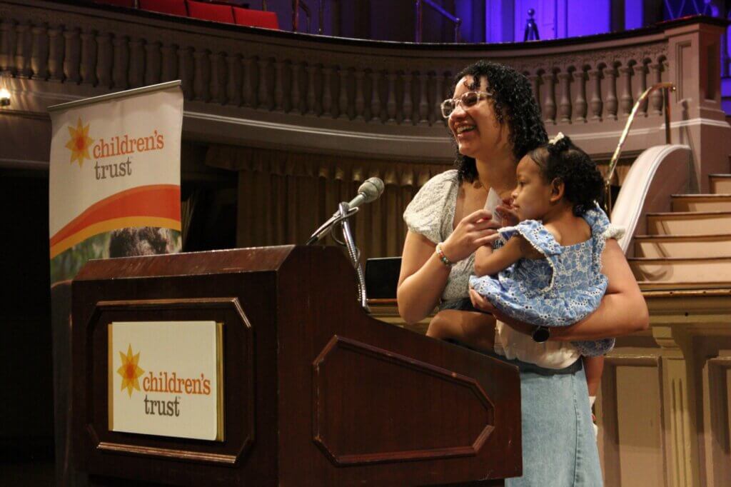 A woman smiles while holding a young child at a podium with a microphone. Behind them, a banner reading Children's trust and an ornate balcony with empty seats set the scene for honoring Massachusetts families at the Celebration Day event.