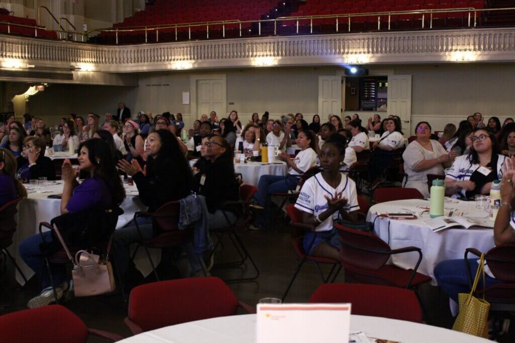 Audience members, mostly women, sit at round tables in a warmly lit hall with red stadium seating above. Many from Massachusetts families are attentively watching and clapping, honoring something special on this Celebration Day.