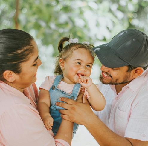 A smiling woman and man are holding a happy toddler between them outdoors. The toddler, in a denim overall and pink shirt, enjoys the playful training moment with parents dressed in light colors, as all three share pure joy together.