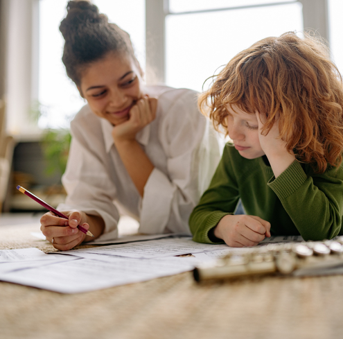 A woman and a child lie on the floor together, looking at sheet music during their training session. The woman smiles and holds a pencil, while the focused child in a green shirt studies. A flute rests in the foreground.