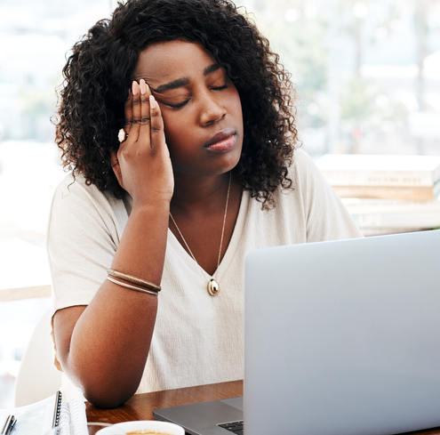 A woman with curly hair sits at a desk in front of an open laptop, touching her temple with her eyes closed as if feeling stressed or overwhelmed during an online training session.