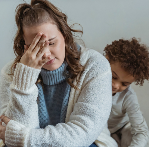 A woman with her hand on her face appears stressed while a child sits behind her, looking down—capturing the emotional challenges of Parental Alienation, and the importance of prioritizing the child's best interest. Both wear cozy sweaters indoors.
