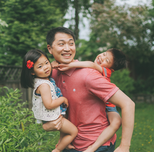 A man smiles while holding two young children in his arms outdoors. The girl, in a white top with a red flower in her hair, and the boy, dressed in red, hug him amid lush greenery.
