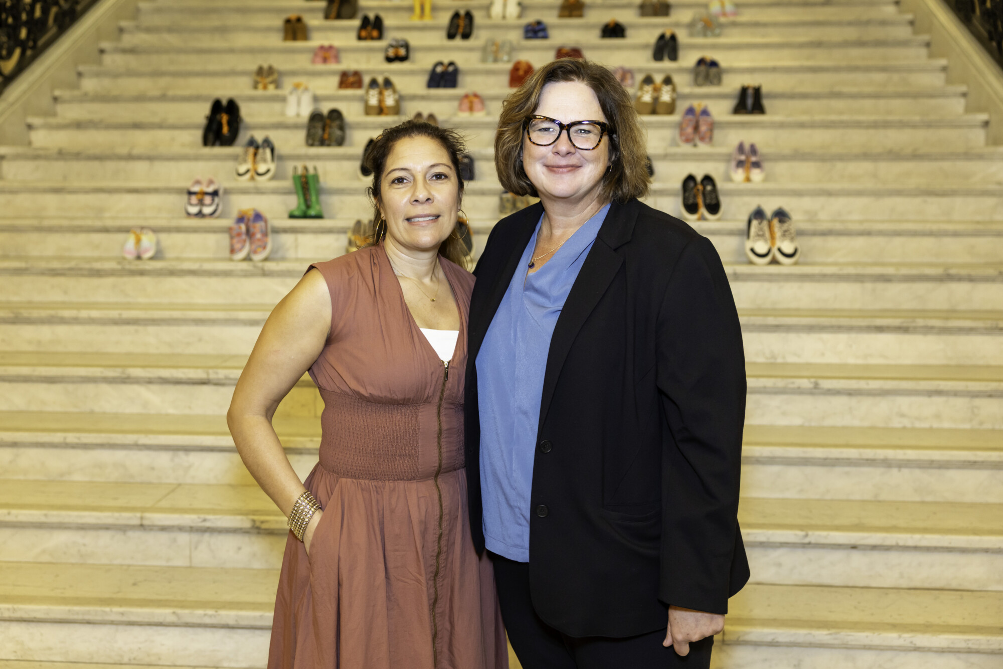 Two women stand together smiling on marble stairs, with many pairs of children's shoes arranged on the steps behind them—a powerful tribute to child sexual abuse prevention in Massachusetts.