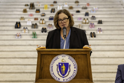 A woman speaks at a podium with the Massachusetts state seal; pairs of shoes arranged on steps behind her symbolize absent individuals, highlighting the urgent need for child sexual abuse prevention in Massachusetts.