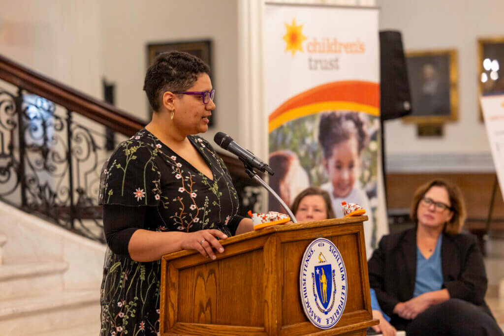 A person speaks at a podium Juanita Cox with two people sitting next to her, including a Children’s Trust banner is displayed in the background near a staircase, highlighting Step up for Kids, Child Abuse Prevention Month.