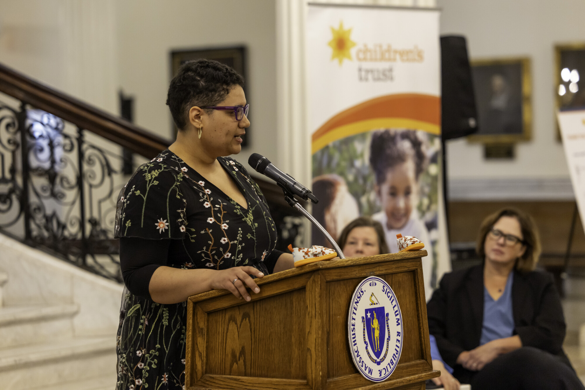 A person speaks at a podium Juanita Cox with two people sitting next to her, including a Children’s Trust banner is displayed in the background near a staircase, highlighting Step up for Kids, Child Abuse Prevention Month.