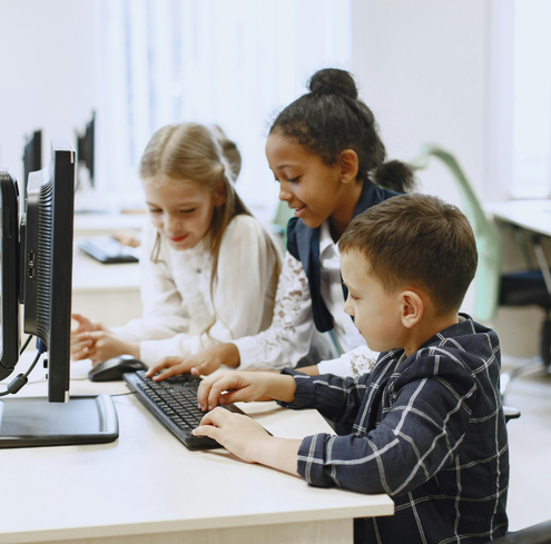 Three young children work together at a desktop computer in a classroom setting, smiling and interacting with the keyboard and monitor.