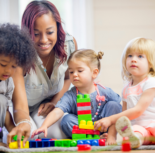 A woman smiles while sitting with three young children playing with colorful building blocks on the floor.