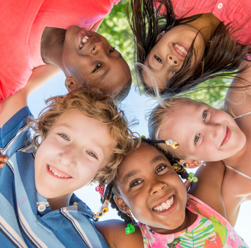 Five smiling children stand close together in a circle, looking down at the camera with their arms around each other, outdoors on a sunny day with green leaves visible in the background.