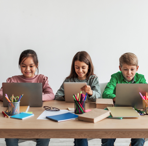 Three children sitting at a table using laptops and smiling. The table has books, papers, and containers with colorful pencils on it. The background is a plain white wall.