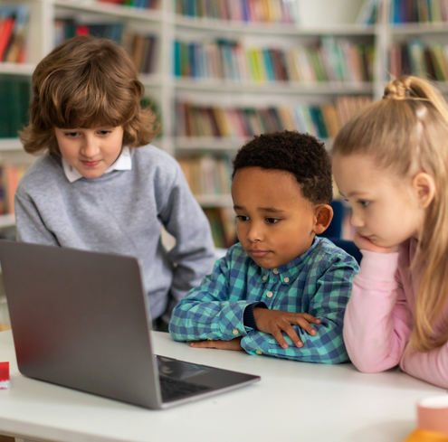 Three young children sit together at a table in a library, watching something on a laptop screen. Bookshelves filled with books are visible in the background.