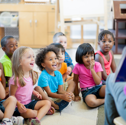 A group of young children sit on the floor in a classroom, smiling and laughing as they listen to an adult who is out of frame.