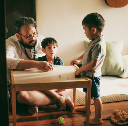 A Father and two young boys sit around a small table playing with toy cars in a cozy room with cushions and a guitar on the wall.
