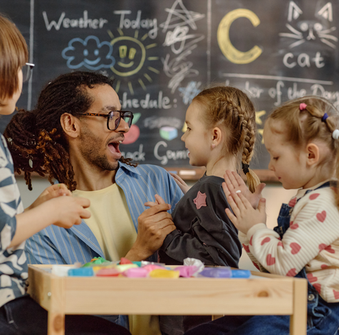 A teacher with glasses and dreadlocks laughs and interacts with three young girls in a classroom. They sit at a table with colorful clay; a chalkboard with drawings and writing is in the background.