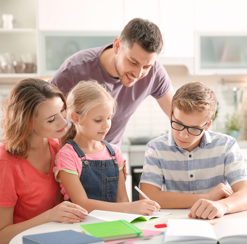 A family of four sits at a table. The mother and daughter look at a notebook, while the father watches and the son writes. School supplies are on the table, and the family appears to be working on homework together, demonstrating teamwork and supportive parenting.