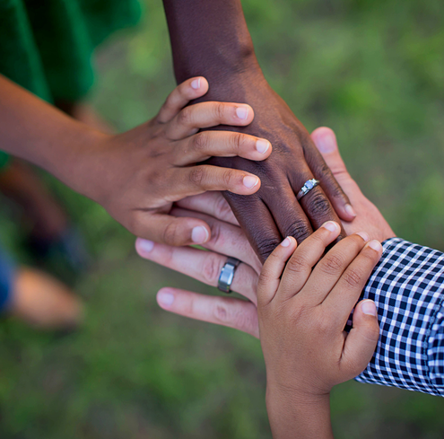Five hands of different skin tones, including adults and children, stack together in a gesture of unity. The outdoor background is blurred with greenery. Several hands have rings, and one person wears a checkered sleeve.