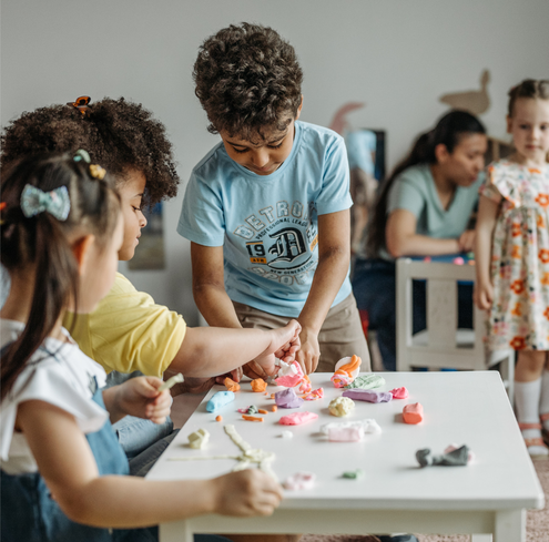 Children gather around a white table, as they play with colorful modeling clay. In the background, a woman sits with another child, guiding them through creative exercises. The scene is indoors and vibrant, with a focus on creativity, interaction, and hands-on learning.