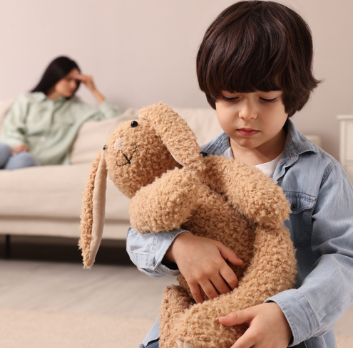 A sad young boy hugs a stuffed bunny while a woman sits on a couch in the background, looking distressed with her hand on her forehead, perhaps struggling.