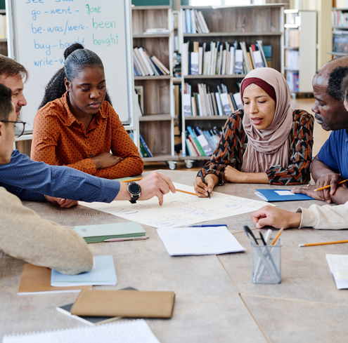 Six adults sit around a table in a library, collaborating and discussing notes on a large sheet of paper with bookshelves and a whiteboard in the background.