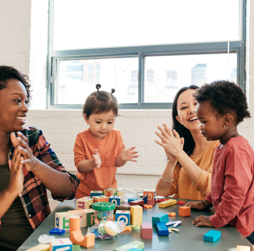 Two adults and two young children with colorful wooden blocks at a table, smiling and clapping together in a bright room with a window in the background.