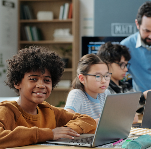 A smiling child in a brown sweater sits at a desk with a laptop in a classroom. Two other children work at laptops nearby, and an adult stands in the background. Bookshelves and a whiteboard are visible.