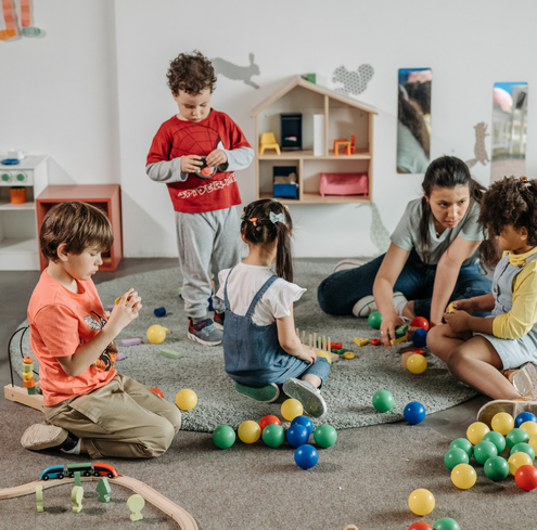 Four children and a woman sit on a rug and floor with colorful plastic balls and toys in a playroom with shelves and wall decorations.