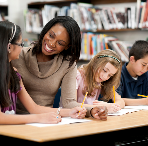 A teacher smiles while providing training to three students with their work at a table in a library or classroom, with bookshelves in the background.