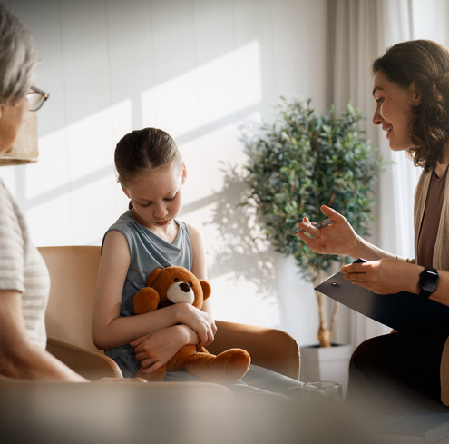 A young girl holding a teddy bear sits beside an older woman while talking to a woman with a clipboard, suggesting a therapy or counseling session in a bright room.