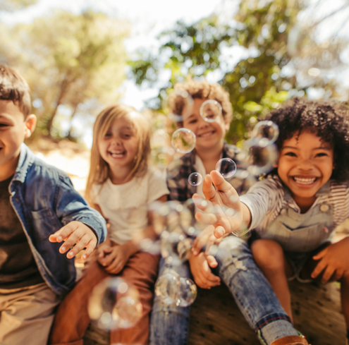 Four children sitting outdoors, smiling and laughing as they reach out to touch floating bubbles with trees and sunlight in the background.