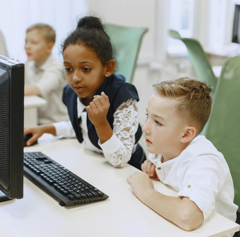 Two children sit at a desk, focused on a computer screen. One girl points at the monitor while a boy watches attentively. Other students are blurred in the background, suggesting a classroom or computer lab setting.