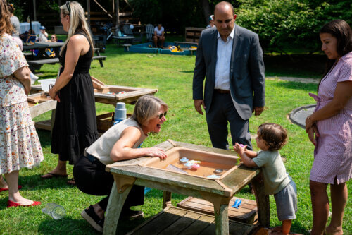 A woman kneels by a water play table, smiling at a toddler. Three adults stand nearby, watching, The scene is outdoors on a sunny day with grass, trees, and other people in the background.