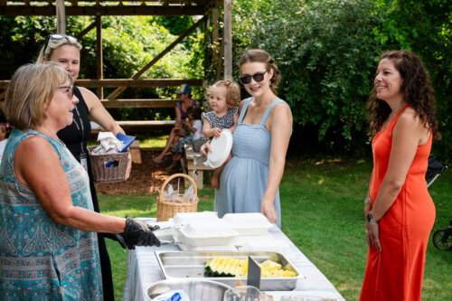 Four women, one holding a toddler, stand outside at a food table with containers of fruit. They are smiling and chatting in a sunny, green garden setting an uplifting scene.