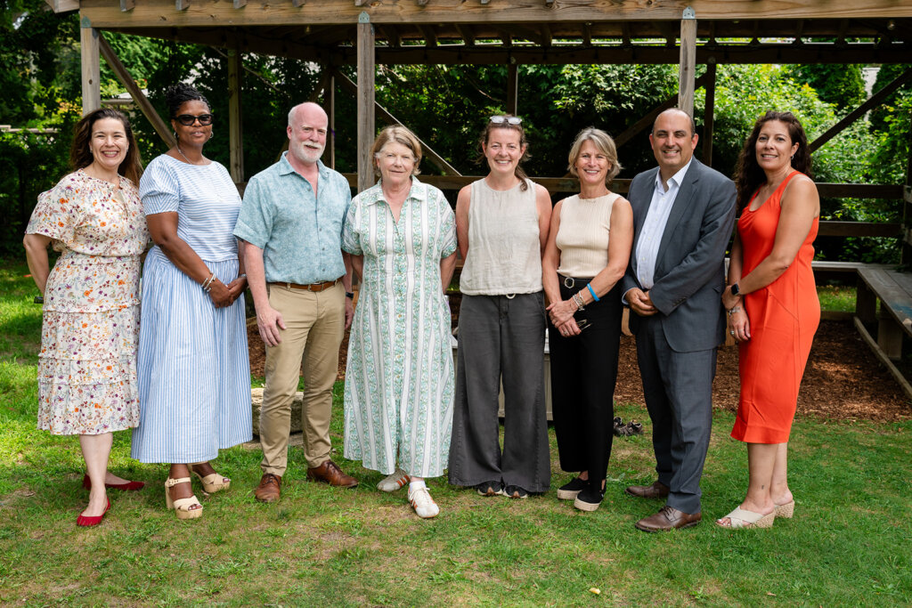 Eight adults stand in a row outside on grass, smiling at the camera. They are dressed in summer clothing and there is a wooden structure with greenery in the background, Martha’s Vineyard Community Services as they gather to support family support policy initiatives.
