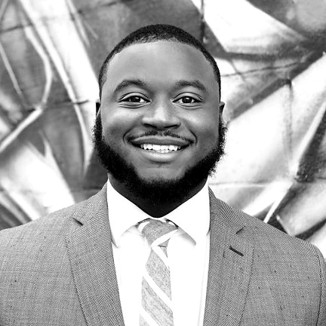 Matthew Kincaid, dressed in a suit and tie, smiles in front of an abstract, geometric mural background. The image is in black and white.
