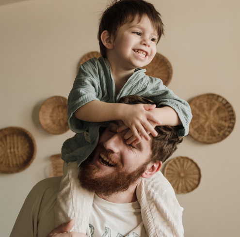 A young child sits on a man’s shoulders, both laughing with their hands touching.