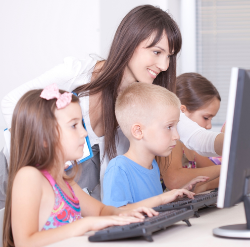 A teacher assists three young students as they use desktop computers to learn about online safety for kids in a classroom.