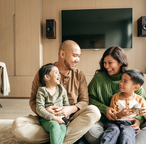 A family of four sits together on a rug in their living room, laughing and playing with small training toys.