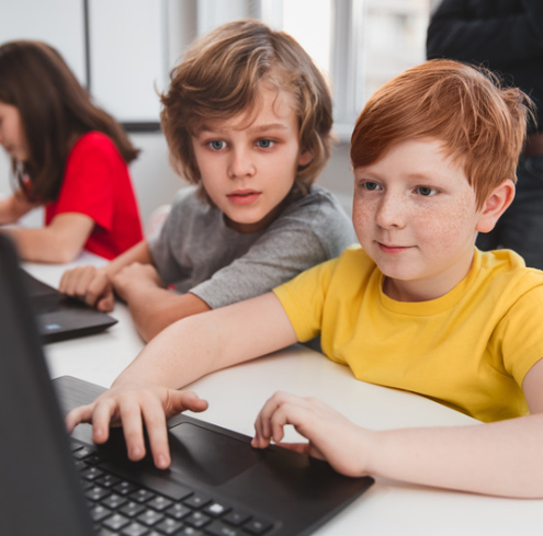 Two children sitting at a desk looking at a laptop computer.