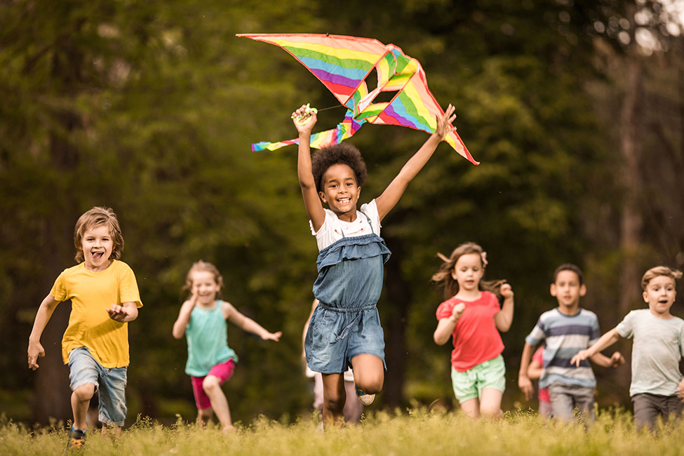 A group of children, run through a grassy field, with one child in the center holding a rainbow kite high above her head.
